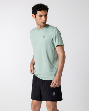 A man wearing a light sage green athletic t-shirt and black athletic shorts, shown from the waist up against a plain light gray studio background.