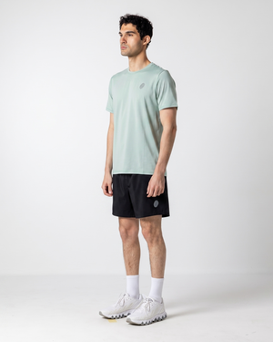 A man wearing a light sage green athletic t-shirt, black athletic shorts, white socks, and white running shoes, standing in profile against a light gray studio background.