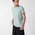 A man wearing a light sage green athletic t-shirt and black athletic shorts, shown from the waist up against a plain light gray studio background.